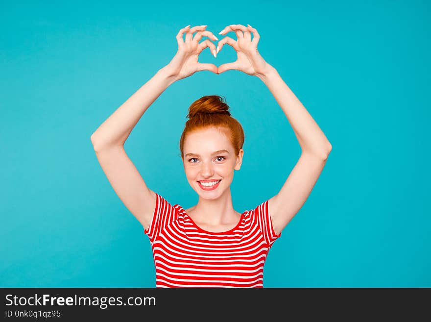 Portrait of nice cute adorable attractive lovely stylish cheerful positive red-haired girl with bun, wearing t-shirt, raising hands up, showing heart, isolated on bright vivid blue background