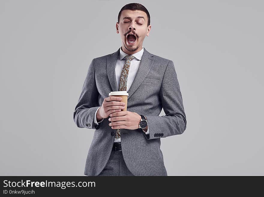 Portrait of handsome young confident arabic businessman with fancy mustache in fashion gray full suit yawns with a cup of coffee on studio background. Portrait of handsome young confident arabic businessman with fancy mustache in fashion gray full suit yawns with a cup of coffee on studio background