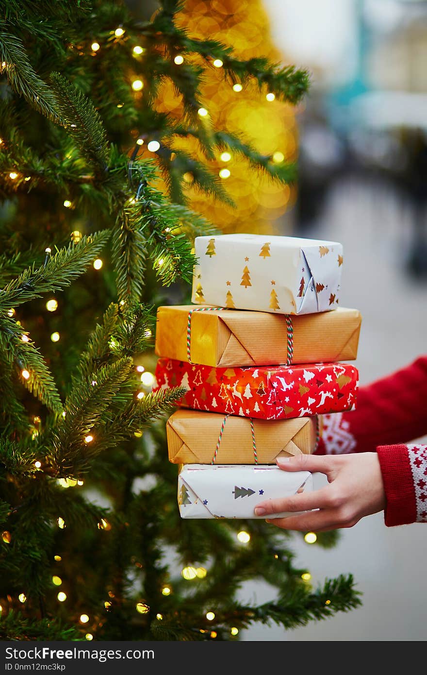 Woman hands holding pile of Christmas presents near New year tree decorated with lights and beads