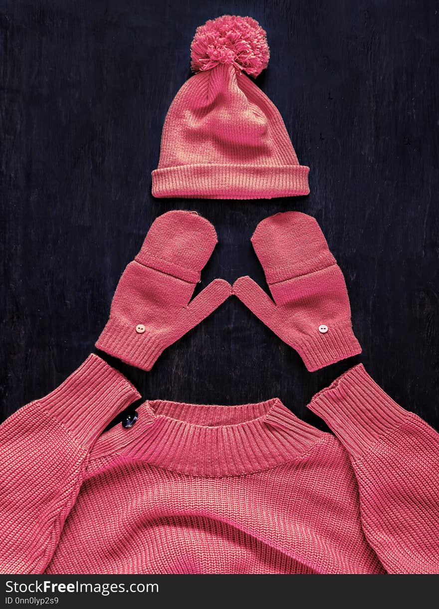 Cap with pompon, mittens, sweater in pink-red-manzhent-purple color on a dark wooden background. Top view, flat lay.Concept stylish youth warm autumn-winter