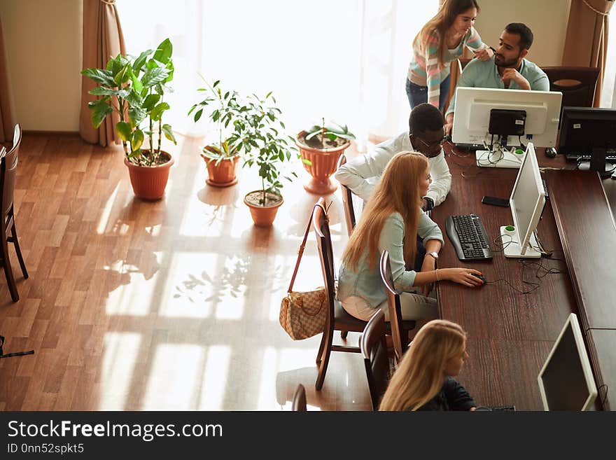 Top view of several people using a personal computer in public place sitting at tables in large spacious well-lit hall with green palnts against sunlight window. Top view of several people using a personal computer in public place sitting at tables in large spacious well-lit hall with green palnts against sunlight window.