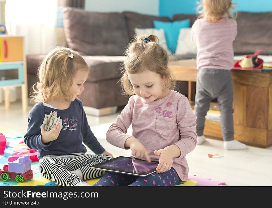 Two little sisters use tablet at home and sit in the middle of toys. Watching something funny