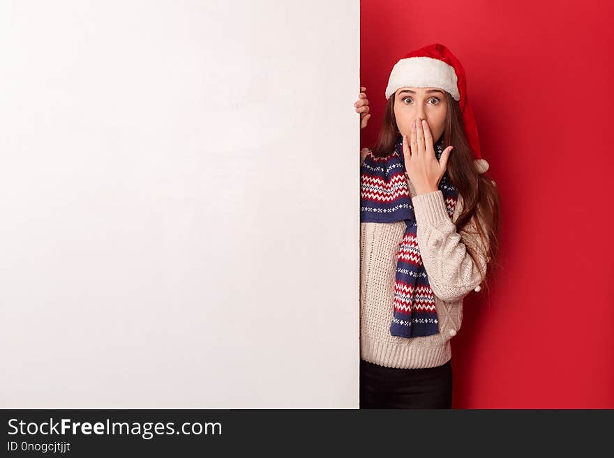 Freestyle. Young woman wearing scarf and santa hat standing isolated on red with white board covering mouth shocked