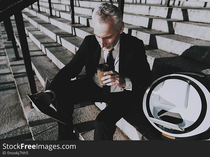 Businessman is Using a Tablet PC. Businessman is Old Smiling Man. Man is Holding a Coffee Cup. Man Wearing in Black Suit. Businessman is Sitting on Staircase. Monowheel near a Man. Sunny Daytime