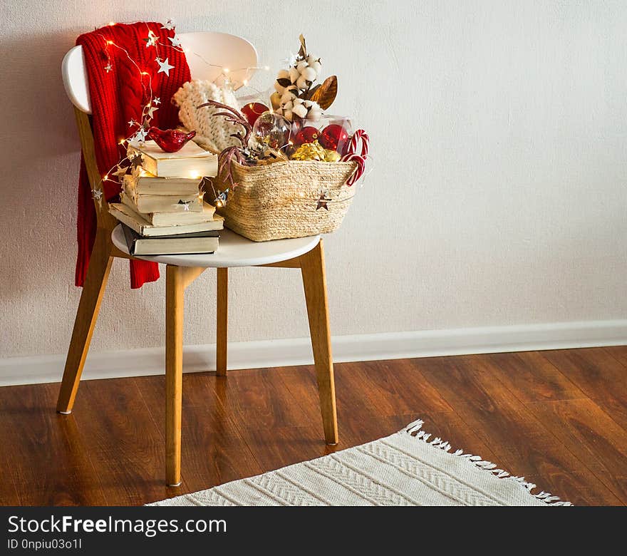 Two winter sweaters laid on a chair with a basket of Christmas decorations, books, led string lights. Winter reading. Winter mood