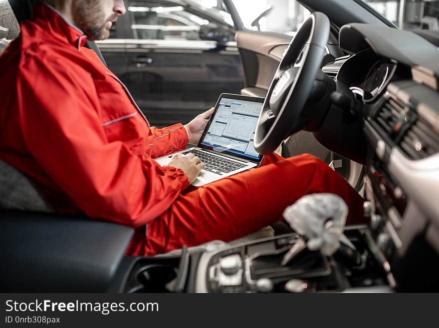 Auto mechanic in red uniform diagnosing car with computer sitting on the driver seat at the car service. Auto mechanic in red uniform diagnosing car with computer sitting on the driver seat at the car service