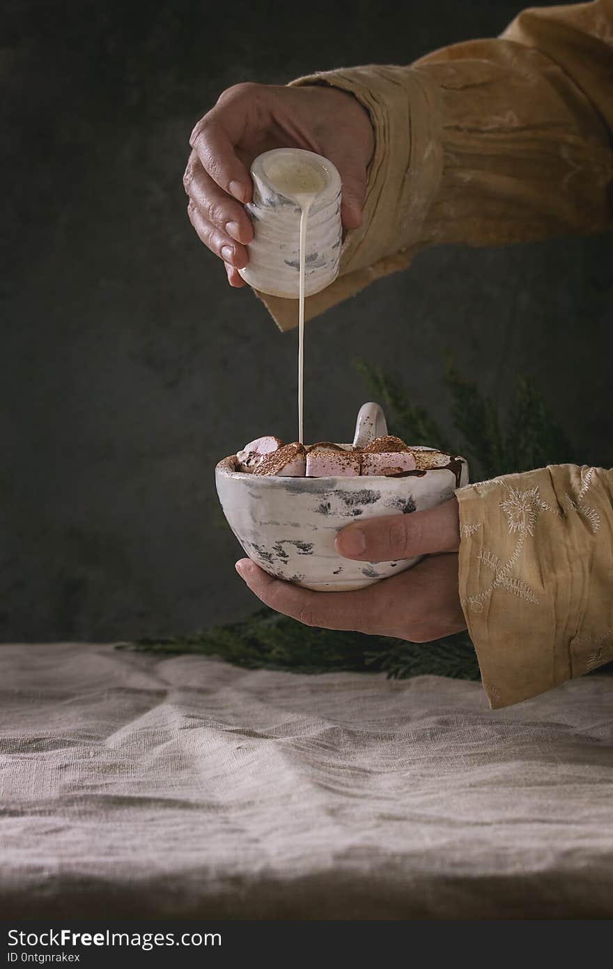 Ceramic cup of hot chocolate with marshmallow s`mores in female hands. Pouring cream from jug. Grey linen table cloth. Winter drink. Dark atmosphere.