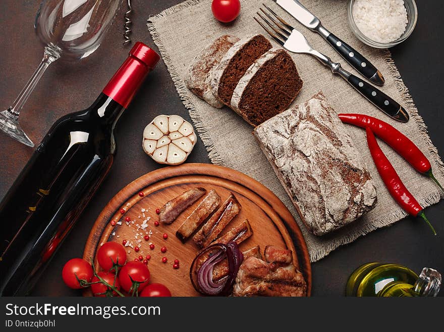 Sliced grilled pork steaks with bottle of wine, wine glass, corkscrew, knife, fork, black bread, cherry tomatoes, garlic, onion and rosemary on rusty background. Top view