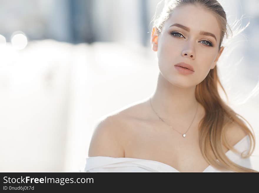 A young beautiful woman with long, hair and gray eyes, beautiful makeup, pink plump lips, sweet smile,blonde hair, spending her time outdoors in the summer in the city, dressed in a white blouse, posing in the sunlight. Portrait of happy young woman. A young beautiful woman with long, hair and gray eyes, beautiful makeup, pink plump lips, sweet smile,blonde hair, spending her time outdoors in the summer in the city, dressed in a white blouse, posing in the sunlight. Portrait of happy young woman