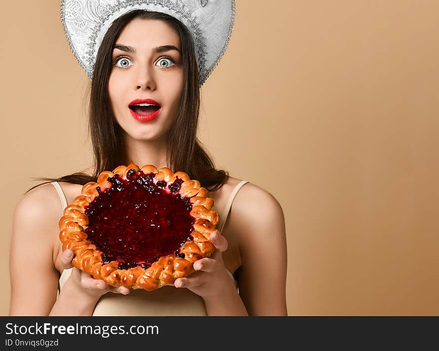 Shocked beauty russian woman in kokoshnik hat, has cake during dinner time, being surprised with its stands against nude beige background. Eating concept