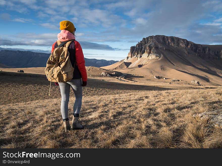 Portrait from the back of a girl traveler in a jacket with a cap and a backpack stands on the background of an epic landscape with rocks.