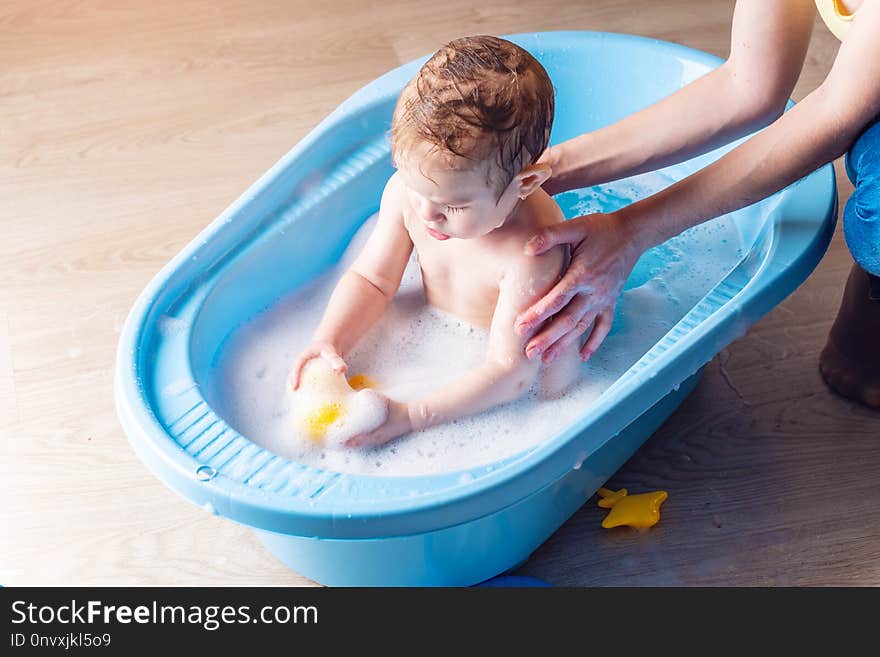 Mom washing little boy in a blue bath in the bathroom. Baby playing with a yellow duck and soap bubbles