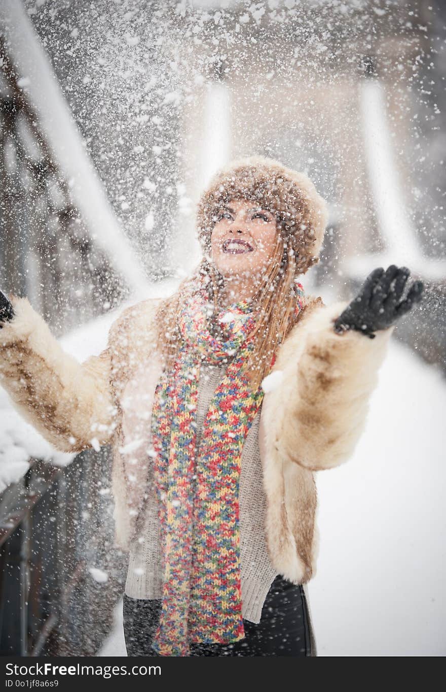 Attractive woman with brown fur cap and jacket enjoying the winter. Side view of fashionable blonde girl posing against snow covered bridge. Beautiful young female with cold weather outfit