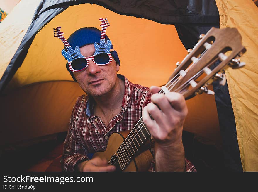 Portrait of joyful hipster man in the funny glasses resting in a tent and playing the ukulele, having fun in travel