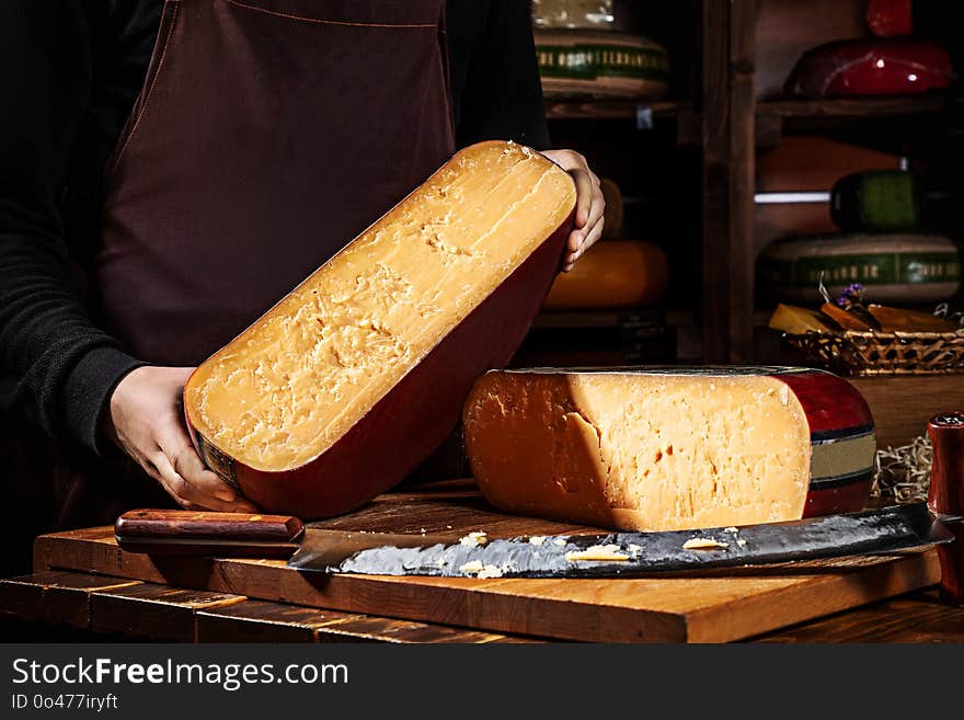 Young worker holding half of wheel gauda cheese in shop. Wooden background, knife for cheese. Close up view. Copy space for text