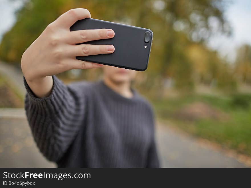 The girl in a gray knitted sweater, takes a selfie on the phone, against the pleasantly blurred trees with autumn, yellow-green foliage and asphalt alley. The girl in a gray knitted sweater, takes a selfie on the phone, against the pleasantly blurred trees with autumn, yellow-green foliage and asphalt alley