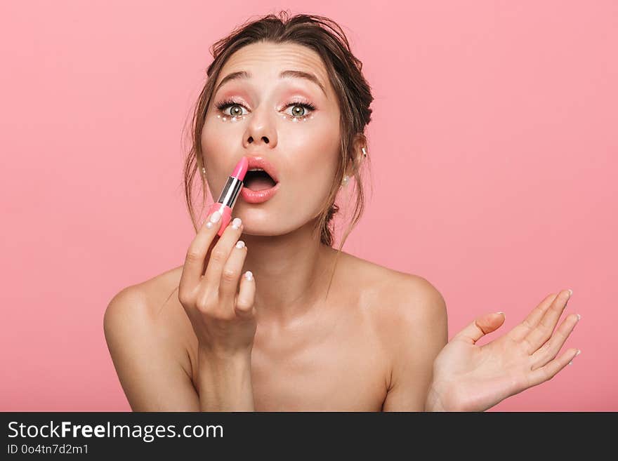 Image of a pretty young woman posing isolated over pink wall background holding lipstick doing makeup