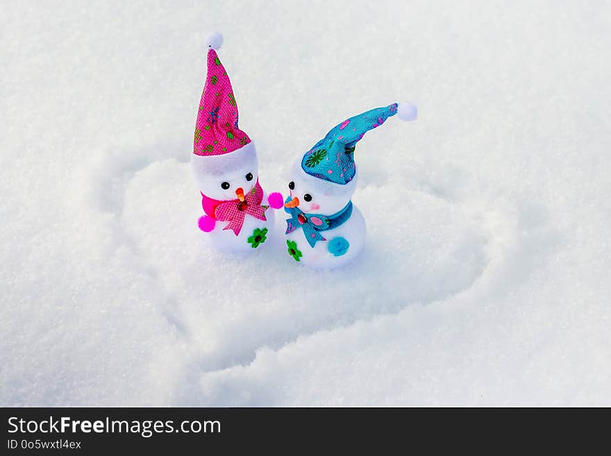 Toy snowmen are in the snow on which the heart is painted. Congratulations on Valentine`s Day_