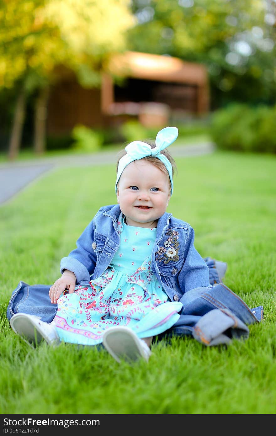 Little happy female child sitting on green grass and wearing jeans jacket.