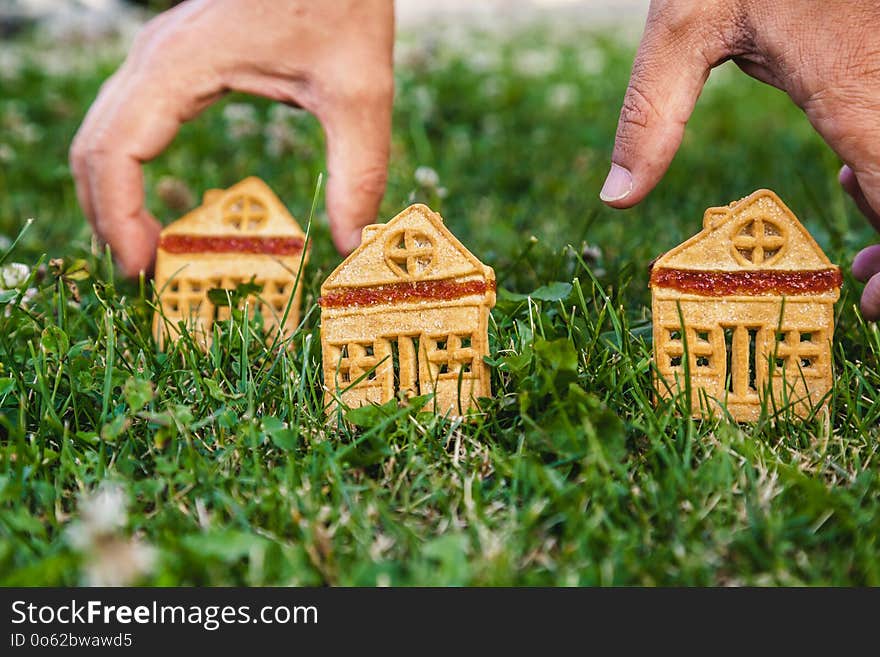 Fabulous sweet house. Three cookies in the form of a house stand on a green meadow close-up. The hand of man puts one house. Fabulous sweet house. Three cookies in the form of a house stand on a green meadow close-up. The hand of man puts one house