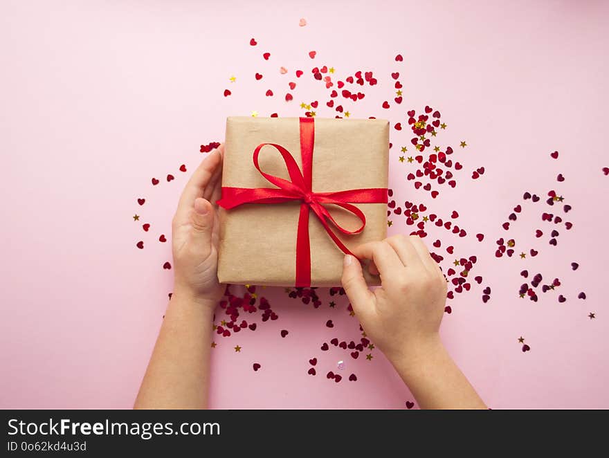 Child is holding gift box wrapped in brown colored craft paper and tied with red bow on pink background with red heart shape confetti. Child is holding gift box wrapped in brown colored craft paper and tied with red bow on pink background with red heart shape confetti.