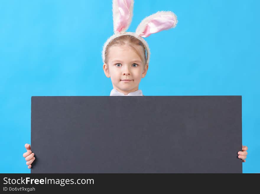 A child in a white rabbit costume on a blue background. A cute little girl with ears of a hare holds in her hands an empty sheet of black cardboard. Advertising photo with space for text.