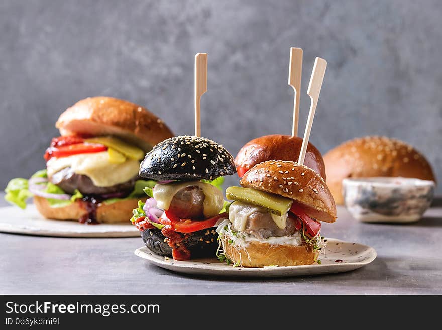 Variety of homemade classic and mini burgers in wheat and black buns with beef and veal cutlets, melted cheese and vegetables on white ceramic board over grey blue table. Variety of homemade classic and mini burgers in wheat and black buns with beef and veal cutlets, melted cheese and vegetables on white ceramic board over grey blue table.