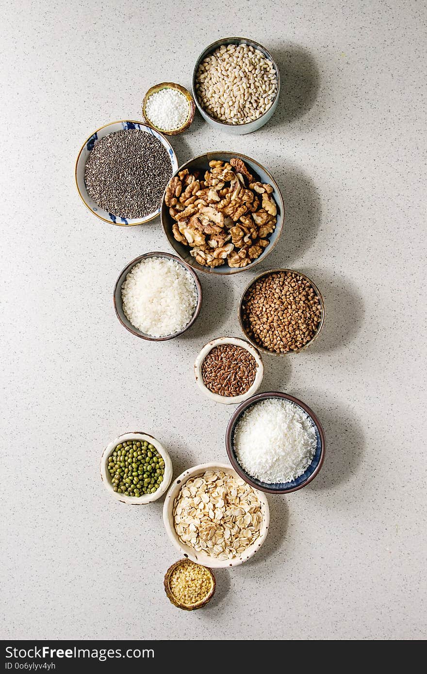 Variety of raw uncooked grains superfood cereal chia, linen, sesame, mung bean, walnuts, tapioca, wheat, buckwheat, oatmeal, coconut, rice in ceramic bowls grey spotted background. Flat lay space. Variety of raw uncooked grains superfood cereal chia, linen, sesame, mung bean, walnuts, tapioca, wheat, buckwheat, oatmeal, coconut, rice in ceramic bowls grey spotted background. Flat lay space