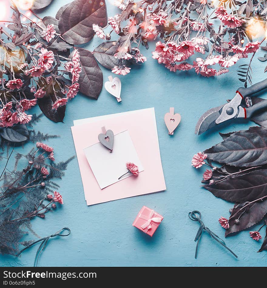 Blank greeting card on blue florist desk with pink flowers, hearts and gift box. Abstract greeting concept. Mothers day, birthday mock up. Copy space. Top view, flat lay