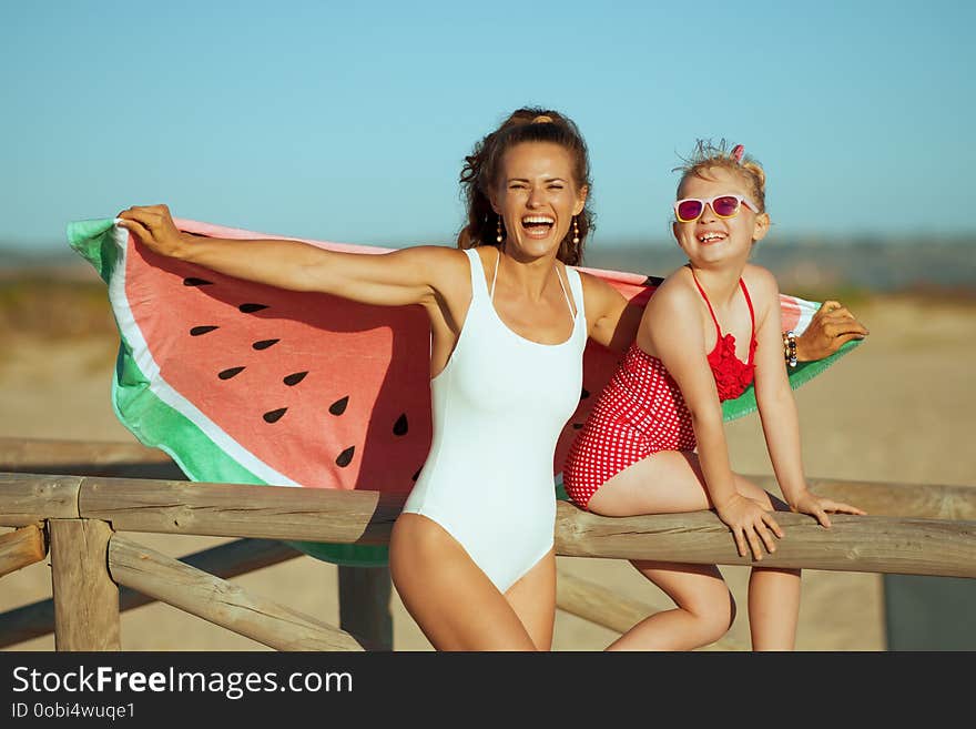 Happy young mother and daughter in swimsuit on the seashore in the evening having fun time holding funny watermelon towel. mother and daughter near a wooden fence. carefree beach fun. Happy young mother and daughter in swimsuit on the seashore in the evening having fun time holding funny watermelon towel. mother and daughter near a wooden fence. carefree beach fun