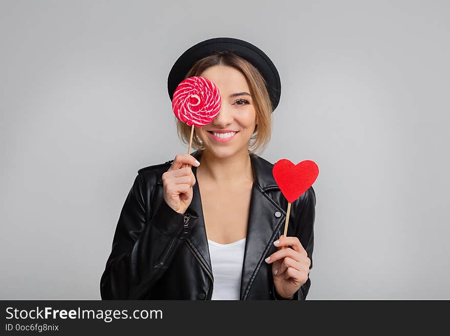 Joyful woman with lollipop and paper heart, having fun, gray studio background