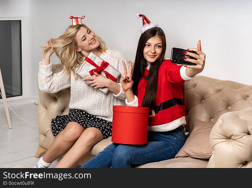 Smiling beautiful young girls in sweaters and santa hats taking a photo of themselves sitting on sofa with gifts. New Year concept.