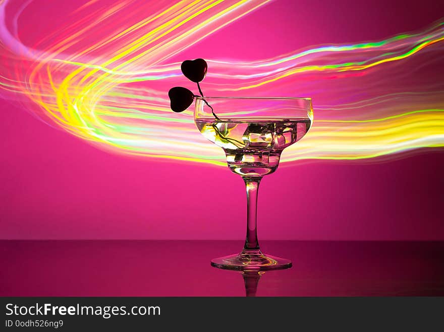 Glass with a cocktail and two hearts. Valentine`s day and wedding concept. On a dark pink background with light highlights