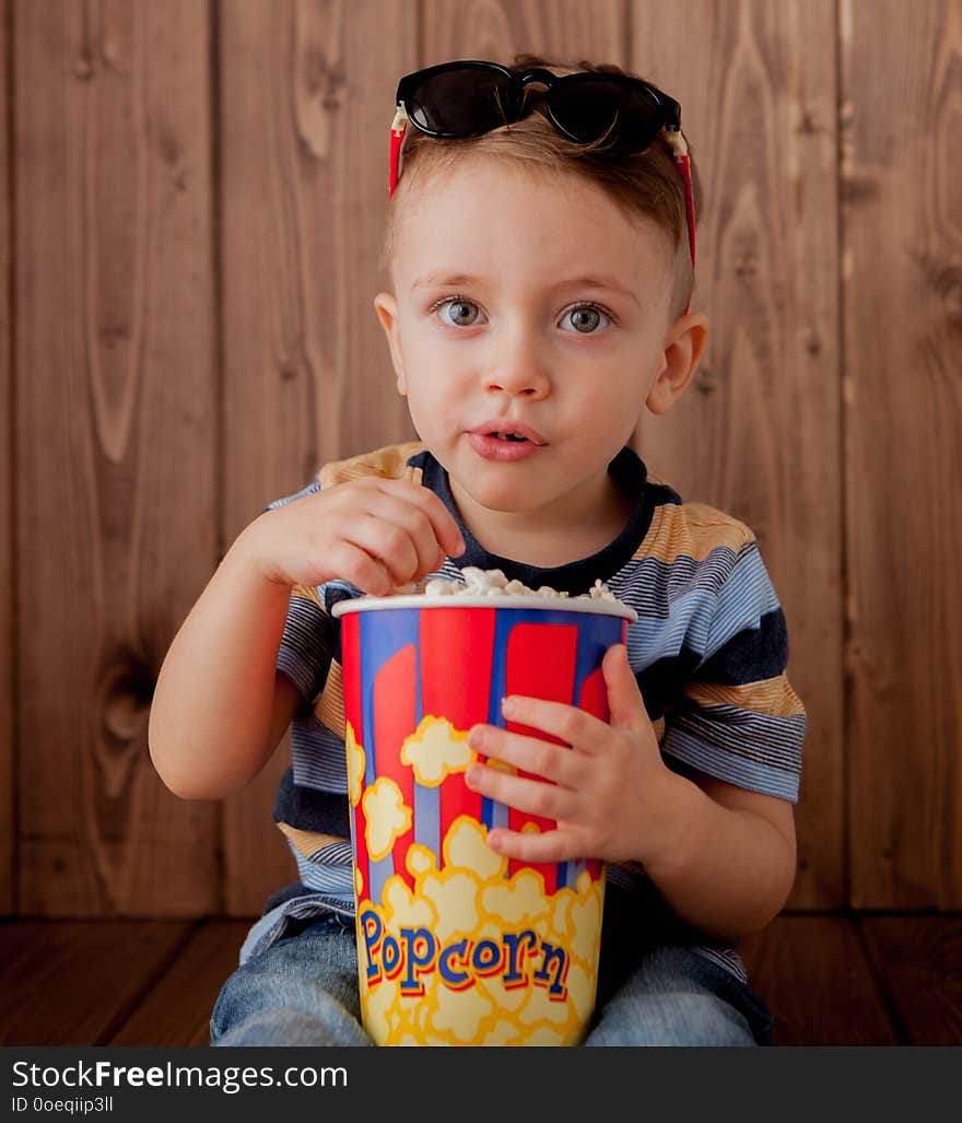 Little cute kid baby boy 2-3 years old , 3d cinema glasses holding bucket for popcorn, eating fast food on wooden background. Kids childhood lifestyle concept. Copy space.