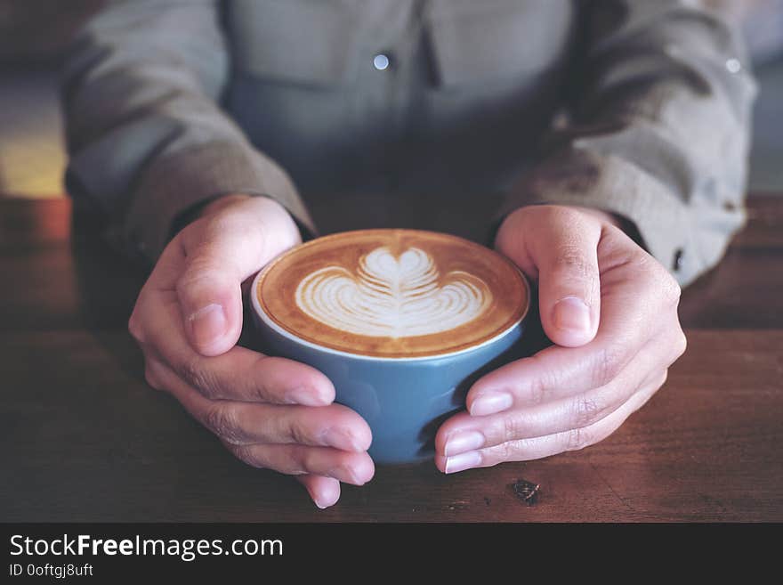 Closeup image of hands holding a blue cup of hot latte coffee with latte art on wooden table in cafe
