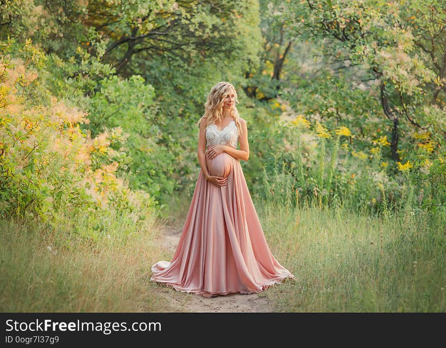 Young sweet pregnant girl with light curly hair hugs and holds her tummy, standing in a fairy forest in cold shades. dresses a long elegant satin silk pink dress with a lace top. art processing photo.