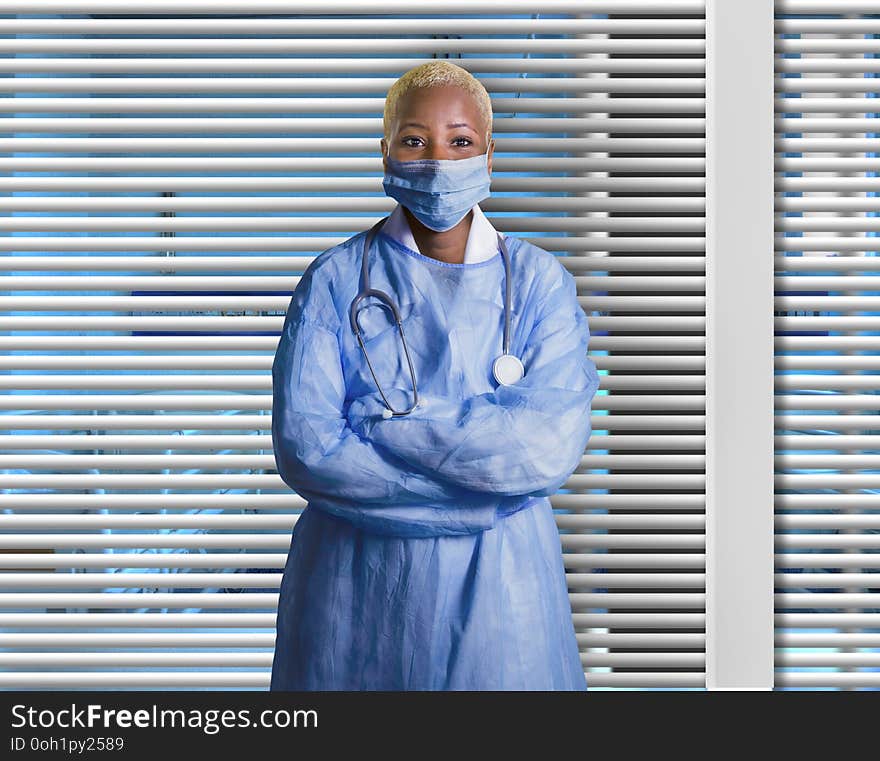 Young attractive and confident black African American medicine doctor wearing face mask and blue scrubs standing corporate at hospital room with Venetian blinds background