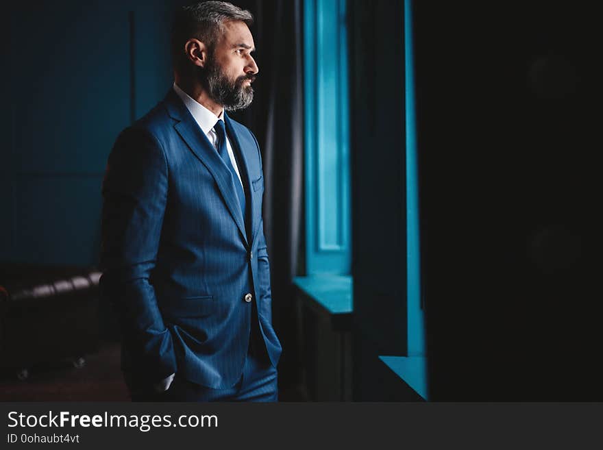 Thinking about... Thoughtful man in full suit looking away through the window while standing indoors.