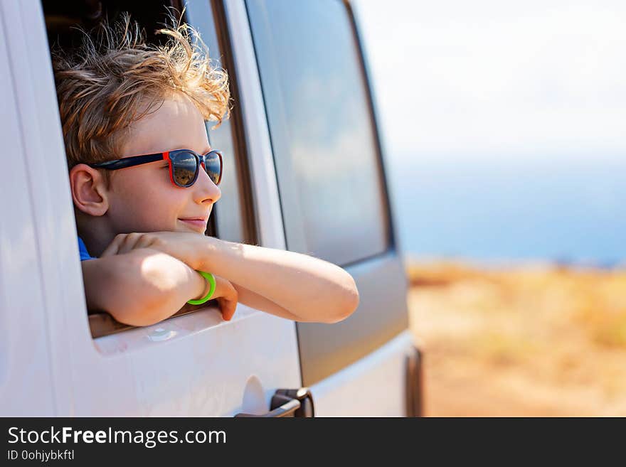 Dreaming positive caucasian boy in sunglasses peeking out of the car window enjoying the ride, tropical family vacation or active road trip concept, copy space on right. Dreaming positive caucasian boy in sunglasses peeking out of the car window enjoying the ride, tropical family vacation or active road trip concept, copy space on right