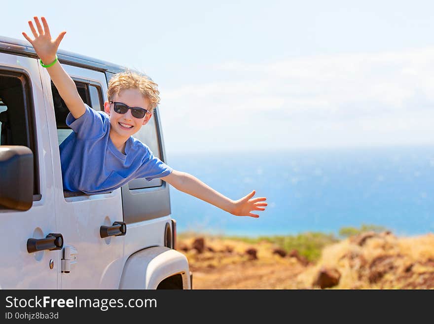 Happy positive caucasian boy in sunglasses peeking out of the car window with his hands up in the air, tropical family vacation or active road trip concept, copy space on right. Happy positive caucasian boy in sunglasses peeking out of the car window with his hands up in the air, tropical family vacation or active road trip concept, copy space on right