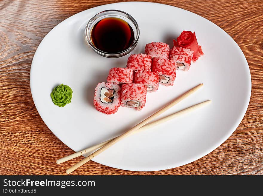 Rolls with salmon and cream cheese, and tomatoes lie on a white plate on which there is still wasabi, pickled ginger, soy sauce and wooden chopsticks, the dish is on a wooden table, flat lay, close up. Rolls with salmon and cream cheese, and tomatoes lie on a white plate on which there is still wasabi, pickled ginger, soy sauce and wooden chopsticks, the dish is on a wooden table, flat lay, close up