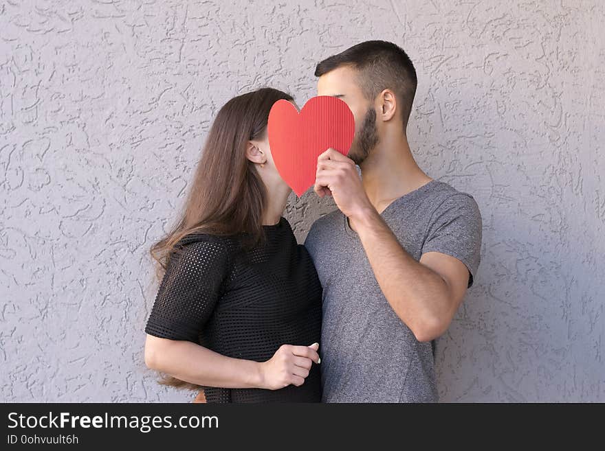 Waist-up portrait of sweet couple deeply in love sharing romantic kiss closing with red paper heart. Intimate moment. Side view. Isolated on grey background. Waist-up portrait of sweet couple deeply in love sharing romantic kiss closing with red paper heart. Intimate moment. Side view. Isolated on grey background