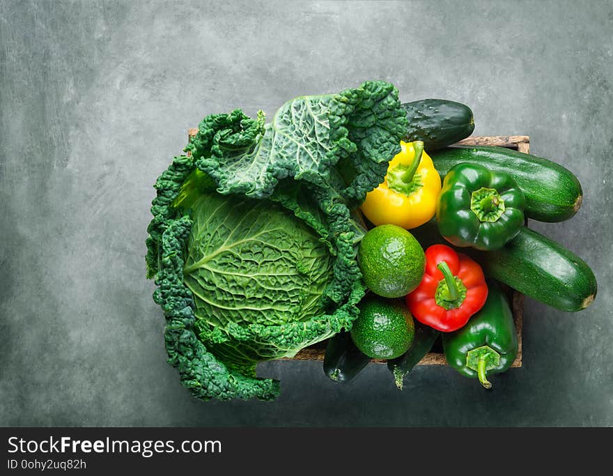 Assortment of organic green red yellow raw vegetables savoy cabbage cucumbers zucchini avocado capsicums in wood box on dark gray concrete background. Healthy lifestyle balanced diet vegan. Top view