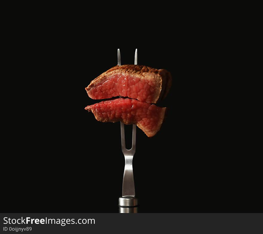 Carving fork with pieces of steak on black background. Tasty meat