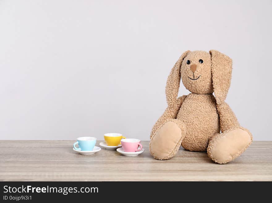 Adorable plush bunny and toy tableware on table against light background, space for text.