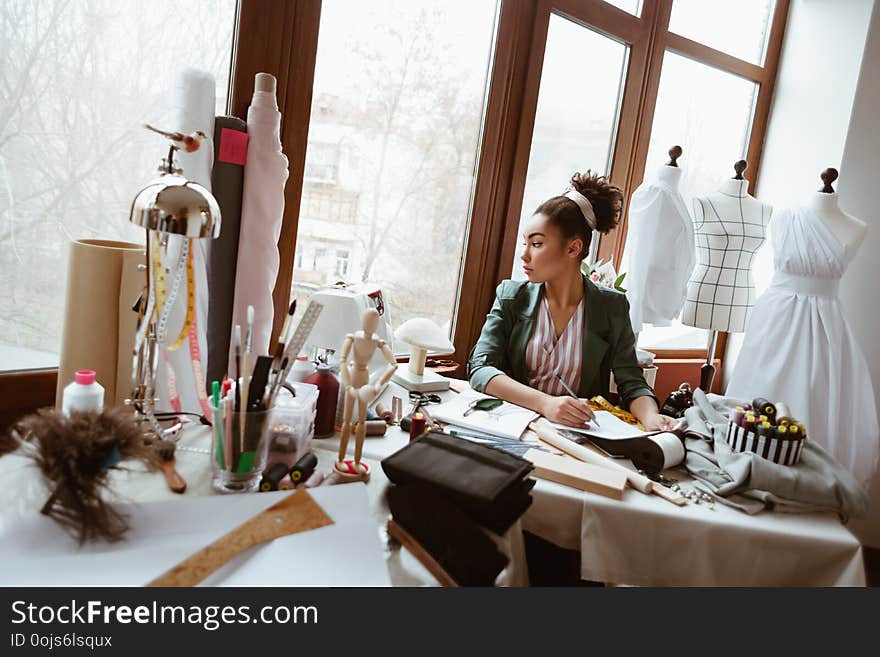 Fashion designer creates new project. Fabulous young woman tailor with sewing machine is thinking about new dress. She is looking to the window thougtful. Fashion designer creates new project. Fabulous young woman tailor with sewing machine is thinking about new dress. She is looking to the window thougtful