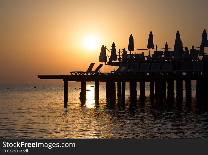 Background with beautiful sunset view of the sea with warm orange and golden hues. There are sun beds and umbrellas on the pier