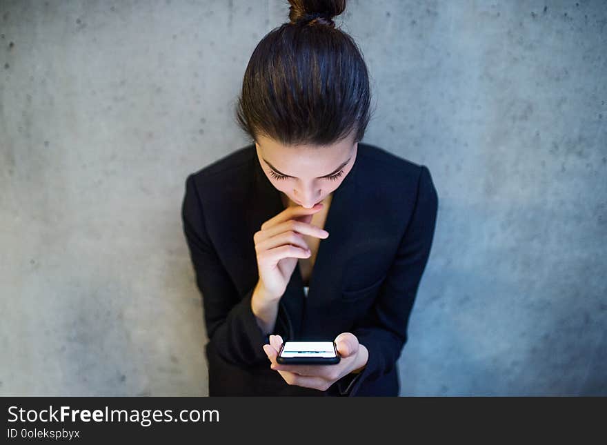 A top view of happy young business woman with smartphone standing against concrete wall in office.