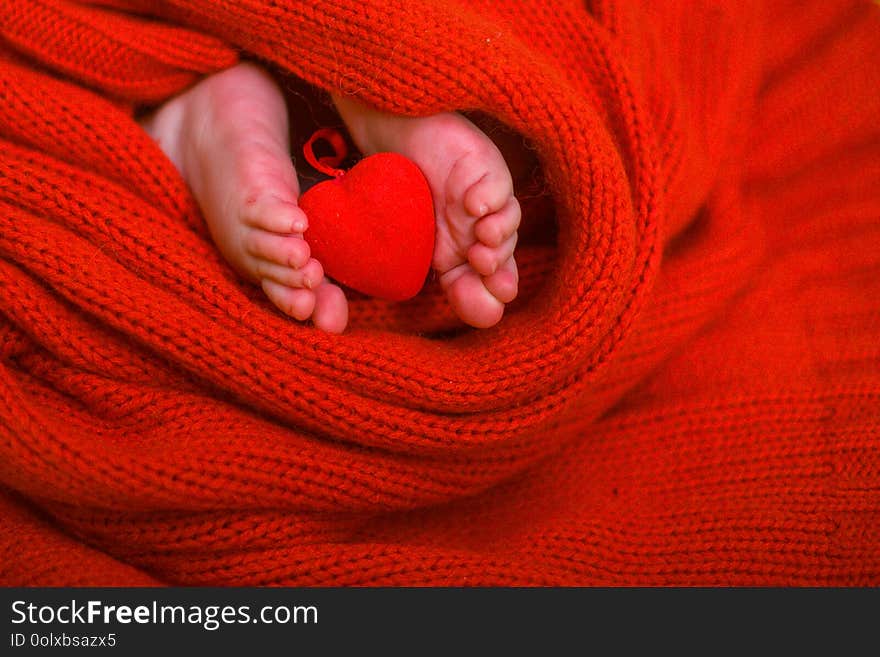 Red heart on baby legs. The legs of the newborn on a red background. A baby wrapped in a red blouse. Valentine`s Day