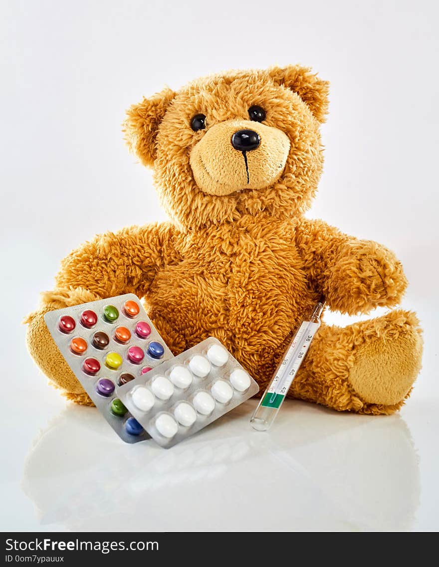 Toy bear with medicine, thermometer and colorful pills in blister pack, sitting on glossy surface against white background. Viewed from its front. Pediatrician office and children health concept